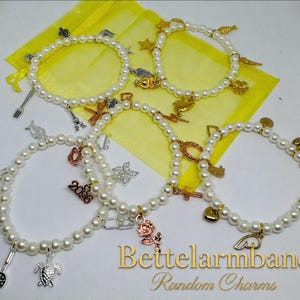 Könnte beinhalten: Vier kreisförmige Armbänder aus weißen Kunstperlen, die jeweils mit einer Auswahl an Metallanhängern in Silber-, Gold- und Roségoldtönen versehen sind. Die Anhänger zeigen Motive wie eine Rose, eine Schildkröte, ein Herz, einen Stern und 2026.
