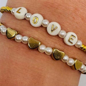 Könnte beinhalten: Zwei Armbänder mit Perlen. Eines zeigt das Wort "LOVE" mit weißen Buchstabenperlen mit goldenen Buchstaben und goldenen Akzenten. Das andere Armband hat goldene herzförmige Perlen und weiße Perlen.