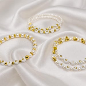 Könnte beinhalten: Drei weiße und goldfarbene Armbänder mit den Wörtern "BRIDE" und "BRIDE SQUAD" in weißen Buchstabenperlen. Die Armbänder liegen auf einer weißen Satin-Oberfläche. Die Armbänder bestehen aus weißen Perlen und goldenen Perlen.