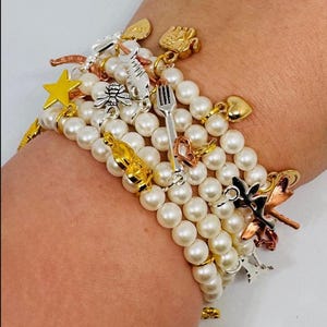 Könnte beinhalten: Ein mehrsträngiges Perlenarmband mit verschiedenen Anhängern. Zu den Anhängern gehören ein goldener Stern, eine Biene, eine Gabel, ein Herz und andere dekorative Elemente. Das Armband wird am Handgelenk getragen.