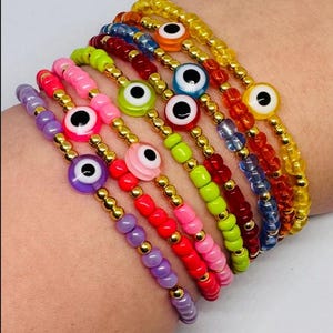 Könnte beinhalten: Eine Auswahl an bunten Perlenarmbändern. Jedes Armband hat einen bösen Blick-Anhänger mit weißer Iris und schwarzer Pupille. Die Armbänder sind in den Farben Lila, Rosa, Rot, Grün, Orange und Gelb gehalten, mit goldfarbenen Perlen.