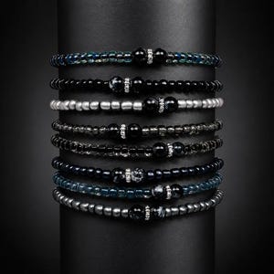 Könnte beinhalten: Eine Sammlung von Armbändern mit Perlen in verschiedenen Farben, darunter Schwarz, Silber und Blau. Jedes Armband hat eine zentrale dekorative Perle mit einem funkelnden Akzent. Die Armbänder werden auf einem schwarzen zylindrischen Ständer präsentiert.