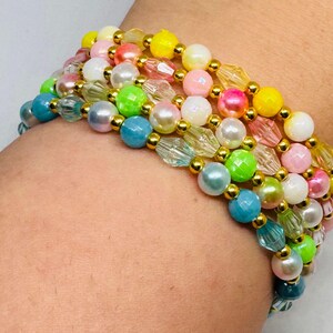 Könnte beinhalten: Mehrere farbenfrohe Perlenarmbänder. Die Armbänder bestehen aus einer Vielzahl von Perlen, darunter runde Perlen, facettierte Kristalle und runde Perlen in Gelb-, Rosa-, Grün-, Blau- und Weißtönen. Goldfarbene Perlen trennen die anderen Perlen.