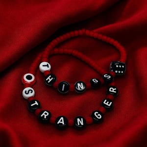 Könnte beinhalten: Zwei Perlenarmbänder aus roten, runden Perlen, durchsetzt mit schwarzen Buchstabenperlen, die STRANGER THINGS bilden. Ein Armband enthält eine weiße Schutzaugenperle mit rotem Zentrum, das andere eine schwarze, sechsseitige Würfelperle.