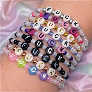 Könnte beinhalten: Ein Stapel aus zehn Perlenarmbändern mit weißen runden Perlen, die mit schwarzen oder goldenen Buchstaben das Wort FUCK bilden. Die Armbänder bestehen aus verschiedenen durchscheinenden, undurchsichtigen und metallischen Perlen in Rosa, Lila, Blau und Silber.