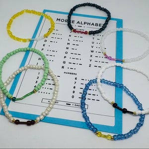 Könnte beinhalten: Sieben kreisförmige Perlenarmbänder in verschiedenen Farben wie Gelb, Schwarz, Hellgrün, Weiß und Blau, die jeweils eine zentrale Stabperle aufweisen. Die Armbänder liegen auf einer weißen Karte mit blauem Rand und einem Morse-Alphabet.