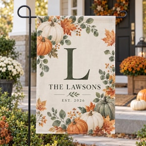 May include: A rectangular garden flag featuring a cream-colored background with a watercolor autumn motif of pumpkins, orange maple leaves, and green foliage. The center displays the letter L, the text THE LAWSONS, and EST. 2026 in dark font.