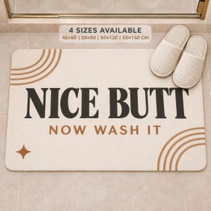 May include: A rectangular beige bath mat featuring the black text NICE BUTT and the brown text NOW WASH IT. The mat includes brown arched line patterns in two corners and a four-pointed star. A pair of white waffle-textured slippers sits on the mat.