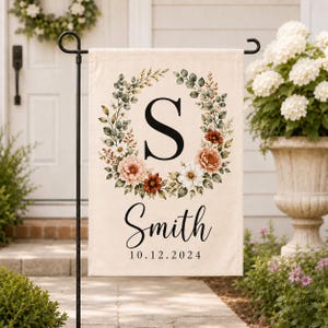 May include: A rectangular garden flag featuring a cream background with a central floral wreath surrounding a large black letter S. Below the wreath, the text Smith is printed in black script, followed by the date 10.12.2024 in black font.