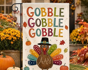 May include: A rectangular garden flag featuring the text GOBBLE GOBBLE GOBBLE in multicolored letters. Below the text is a brown turkey wearing a black pilgrim hat with a yellow buckle. The turkey has colorful, polka-dotted tail feathers.
