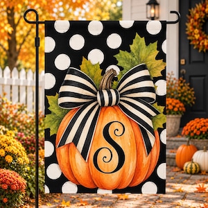 May include: A rectangular garden flag featuring a black background with white polka dots. The center displays an orange pumpkin with a large black and white striped bow and green maple leaves. A black letter S is printed on the pumpkin.