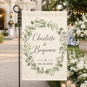 May include: A rectangular off-white garden flag featuring a printed eucalyptus wreath design in shades of green. The center displays the names Charlotte & Benjamin in black script, followed by a small heart icon and the date 06.28.2025.