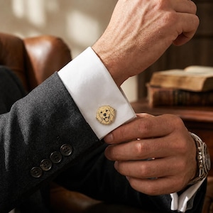May include: A gold-toned metal cufflink featuring a detailed, engraved illustration of a Golden Retriever dog face. The cufflink is attached to the crisp white cuff of a long-sleeved shirt worn under a dark grey textured suit jacket sleeve.