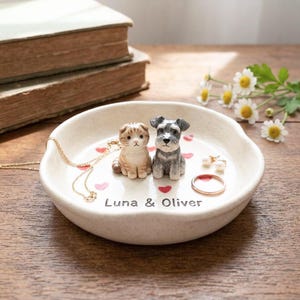 Puede incluir: Un plato de cerámica color crema para anillos con una figura de gato y perro. El plato tiene detalles de corazones rojos y los nombres "Luna & Oliver" impresos. También hay un collar, un anillo y pendientes dorados.