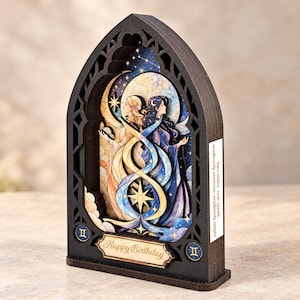 Op de afbeelding: Een gelaagde houten schaduwdoos met een gotisch boogframe met twee figuren die het sterrenbeeld Tweelingen voorstellen. Het kunstwerk bevat hemelse motieven, sterren en manen. Een plaquette toont "Happy Birthday" en de tekst aan de zijkant luidt "Two sides, one spark, endlessly curious, endlessly bright."