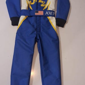Puede incluir: Un traje de carreras azul y blanco con un emblema de león amarillo en el pecho, puños negros y un cinturón con una bandera de los Estados Unidos y el nombre JOEY impreso en blanco. El traje incluye el texto KI y Liga en la parte frontal.
