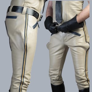 Op de afbeelding: Beige leren broek met zwarte en gouden zijstrepen, een zwarte riem en zwarte laarzen. De broek heeft een klassiek ontwerp met een getailleerde pasvorm. De outfit bevat een bijpassende leren top en zwarte handschoenen.