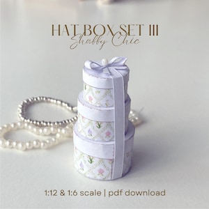 Puede incluir: Un conjunto de tres sombrereras redondas, atadas con una cinta y un lazo color lavanda. Las cajas son blancas con un estampado floral y el texto "HAT BOX SET III Shabby Chic" está en la parte superior. La escala es 1:12 y 1:6.