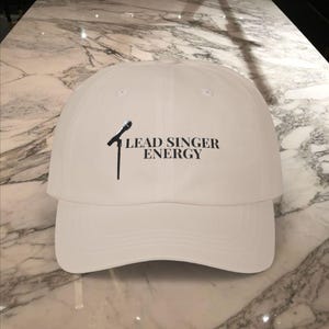 May include: A white baseball cap with a curved peak. The front of the cap displays a black microphone graphic and the words "LEAD SINGER ENERGY" in black. The cap is made of a smooth material.