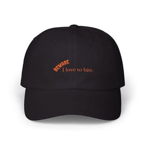 May include: Black baseball cap with a curved brim. The cap features the orange text "BEWARE" above the phrase "I love to bite." The hat is a classic six-panel design, suitable for casual wear.