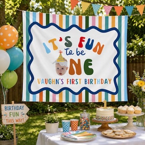 May include: A rectangular birthday banner featuring vertical stripes in blue, orange, green, and pink. The centre displays the text IT'S FUN to be ONE with a photo of a baby wearing a party hat, and the text VAUGHN'S FIRST BIRTHDAY below.
