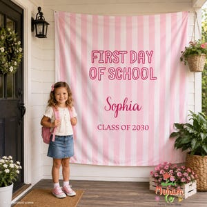 May include: A large fabric backdrop featuring vertical light pink and white stripes with the text FIRST DAY OF SCHOOL, the name Sophia, and the text CLASS OF 2030 printed in a dark pink font, designed for a school-themed photo display.