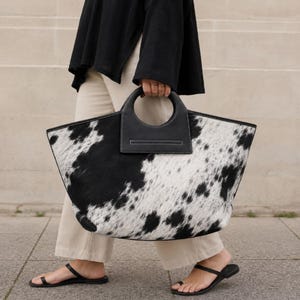 May include: A large tote bag featuring a black and white cow print pattern with a textured hair-on-hide finish. The bag has a wide, trapezoidal shape and a structured black leather handle with a circular cutout design for carrying by hand.