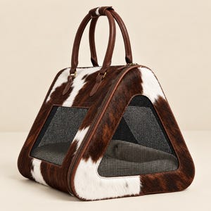May include: A triangular pet carrier featuring a brown and white cowhide pattern. The carrier includes two brown leather handles at the top, a zippered closure, and black mesh ventilation panels on the sides for visibility and airflow.