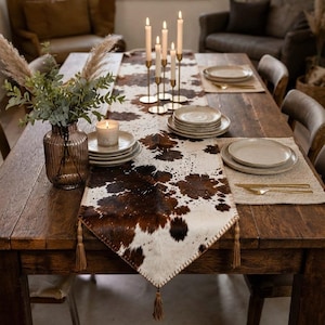 May include: A brown and white cowhide table runner with stitched edges and tassels, displayed on a wooden dining table. The runner is decorated with a glass vase containing dried foliage, a candle, and gold-toned candle holders with white candles.