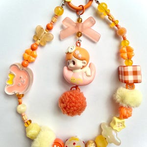 May include: A whimsical orange and yellow beaded charm with a variety of decorative elements. Features include a small figurine, a bow, a pom-pom, and various beads and charms. The charm is attached to a circular orange clasp.