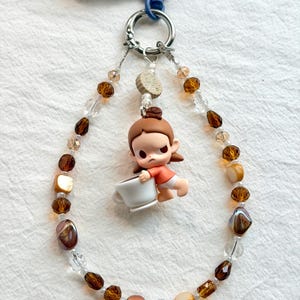 Puede incluir: Un colgante para teléfono con un bucle de cuentas marrones, ámbar y transparentes unido a un anillo de metal plateado. Una pequeña figura de una persona con cabello castaño, camisa naranja y pantalones cortos blancos sostiene una taza blanca.