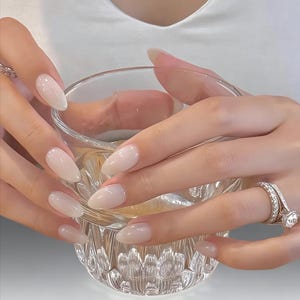 Puede incluir: Primer plano de manos con uñas largas, ovaladas, de color rosa claro que sostienen un vaso transparente facetado. Las manos están adornadas con anillos, uno con una piedra grande y transparente. El fondo es una parte superior blanca.