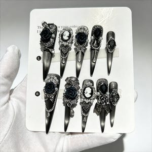 Könnte beinhalten: Ein Set aus zehn stilettoförmigen Press-on-Nägeln in metallischem Anthrazitgrau. Die Nägel sind mit schwarzen 3D-Rosen, weißen Kamee-Porträts, silberfarbenen Metallanhängern, kleinen Kunstperlen und klaren Strasssteinen auf einer weißen Karte.