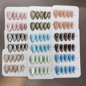 Puede incluir: Una colección de uñas postizas en forma de almendra dispuestas en tarjetas de presentación blancas. El set incluye varios colores como rosa, oro, plata, azul, verde y marrón, con un acabado brillante de efecto ojo de gato.