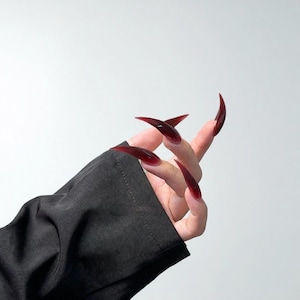 Puede incluir: Un juego de uñas postizas largas y puntiagudas en forma de estilete de color rojo burdeos profundo y translúcido. Las uñas tienen un acabado brillante y se muestran en los dedos, con la mano parcialmente cubierta por una prenda negra.