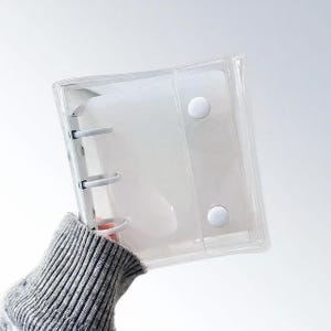 May include: Clear, transparent binder with white details. The binder features a ring mechanism and two white snap closures. The binder is held by a person wearing a grey sweater.