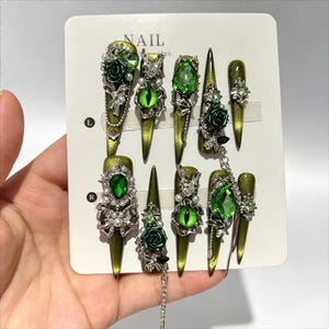 Op de afbeelding: Een set van tien stiletto-vormige press-on nagels in een metallic olijfgroene afwerking. De nagels zijn versierd met zilverkleurige bedels, waaronder driedimensionale rozen, groene strass-steentjes, konijnmotieven en bungelende kettinkjes.