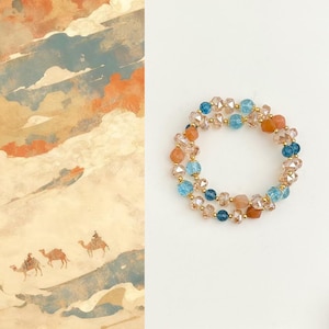 May include: A beaded bracelet with a mix of peach, light blue, and clear crystal beads. The bracelet is strung on a gold-colored wire. The background features a painting of a desert scene with camels.