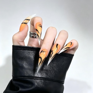 May include: Stiletto-shaped press-on nails featuring a vibrant orange base with black tiger-stripe patterns and white tips. The nails have a glossy finish and are displayed on fingers, with one finger wearing a silver-toned metal ring with a star cutout.