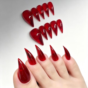Pode incluir: Uma coleção de unhas acrílicas vermelhas, translúcidas e pontiagudas. As unhas estão dispostas em fileiras, algumas exibidas em uma mão. As unhas têm um acabamento brilhante e uma forma de estilete afiada, ideal para um visual ousado.