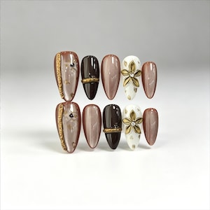 Puede incluir: Un juego de diez uñas postizas en forma de almendra con diseños en marrón oscuro brillante, malva metálico con efecto ojo de gato y base blanca. Incluye adornos de tiras doradas texturizadas, bandas horizontales y flores en relieve.