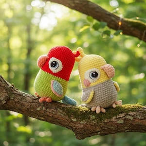 May include: Two handmade crochet bird toys perched on a tree branch. One bird is red and green, the other is yellow and gray. Both have large eyes and are made of yarn. The birds are sitting on a tree branch with green foliage in the background.