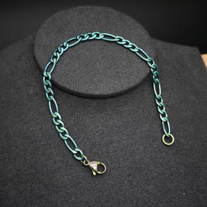 May include: A teal-colored Figaro chain bracelet. The bracelet features a series of elongated and circular links, with a lobster clasp closure. The bracelet is displayed on a dark gray surface.