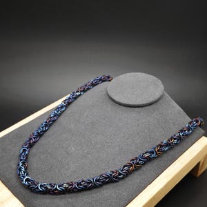 May include: A chain necklace constructed from interlocking metal rings in a Byzantine weave pattern. The rings feature a metallic finish with iridescent hues of deep blue, purple, and copper, creating a multi-colored, reflective appearance.