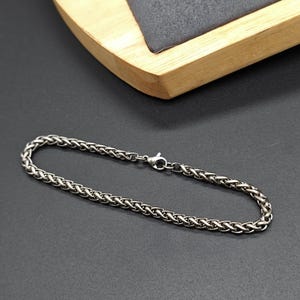 May include: A silver-colored bracelet with a wheat chain design. The bracelet is made of metal and has a lobster clasp closure. The jewelry is displayed on a dark gray surface with a wooden tray in the background.