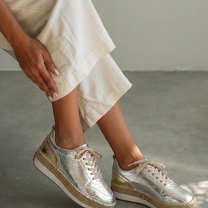 May include: Silver metallic trainers with a perforated design and beige laces. The shoes have a white sole and a woven jute-style trim. The trainers are shown with beige trousers.