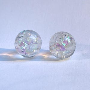 May include: Two clear, round stud earrings with iridescent, glitter-like inclusions. The earrings are set against a white background, creating a minimalist aesthetic. The inclusions reflect light, displaying shades of purple and silver.