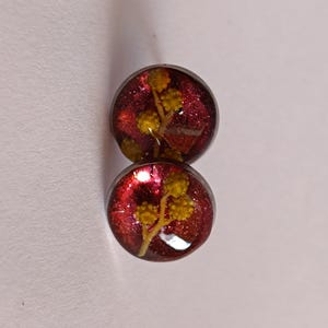 Könnte beinhalten: Zwei runde Ohrstecker. Jeder Ohrring zeigt einen Zweig gelber Blüten, der in klarem Harz mit einem glitzernden roten Hintergrund eingeschlossen ist. Die Ohrringe sind vor einem weißen Hintergrund platziert.