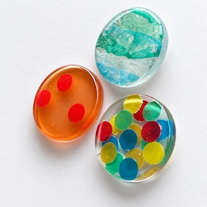 Puede incluir: Tres cabujones de resina circulares. Uno es naranja translúcido con tres puntos rojos. Otro es transparente con papel de aluminio arrugado en color verde azulado y blanco. El tercero es transparente con puntos de confeti de colores.