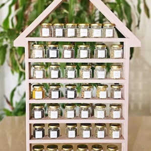 May include: A pink, house-shaped spice rack filled with small glass jars. Each jar has a gold lid and a white label. The rack is made of wood and has multiple shelves. The jars contain various spices, herbs, and seasonings.